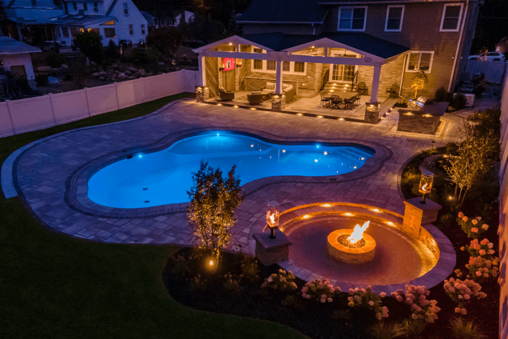 Ground-level nighttime view of the freeform pool with blue LED illumination, raised spillover spa, and fire pit feature