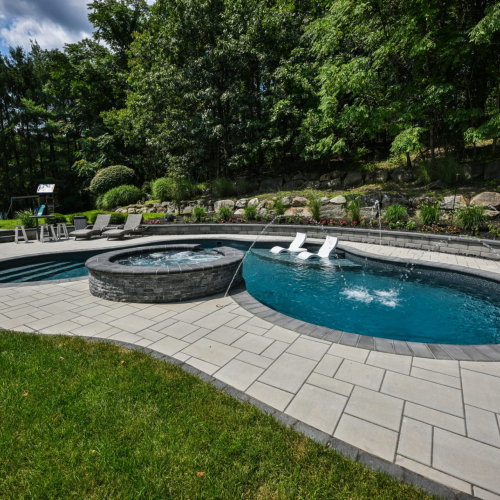 Elegant backyard pool with spa featuring stone accents, surrounded by landscaped greenery and patio seating, showcasing a completed project by EverClear Pools & Spas in Mahwah, NJ.
