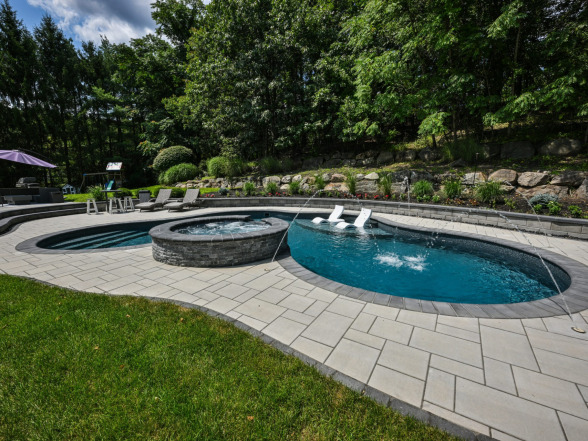 Elegant backyard pool with spa featuring stone accents, surrounded by landscaped greenery and patio seating, showcasing a completed project by EverClear Pools & Spas in Mahwah, NJ.