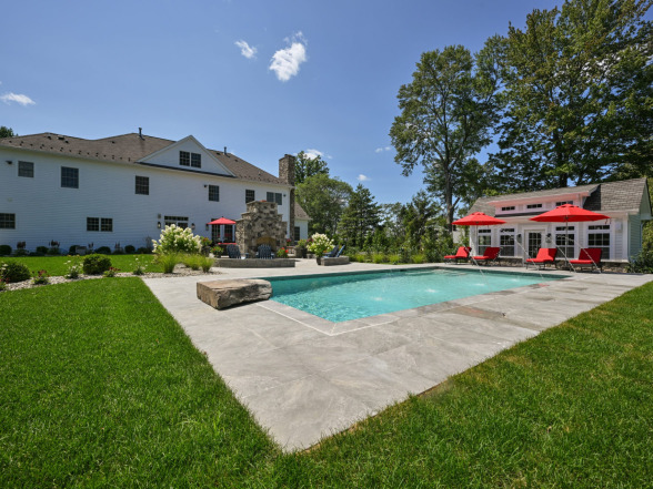 Backyard pool installation by EverClear Pools & Spas, featuring a crystal-clear pool surrounded by lush green grass, stone accents, and patio furniture with red umbrellas, set against a sunny sky and elegant home backdrop.