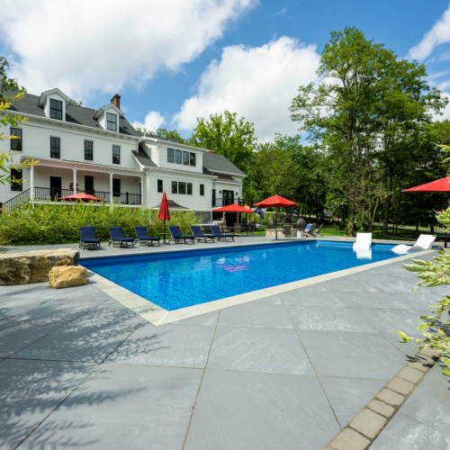 Modern in-ground pool surrounded by a spacious patio, lounge chairs, and red umbrellas, with a stylish white house and lush greenery in Morristown, NJ, showcasing EverClear Pools & Spas pool design and installation services.