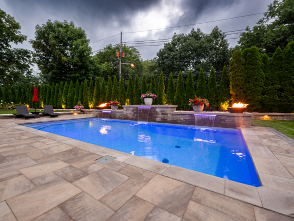 Angled view of the pool highlighting the natural stone paver patio, fire bowls, and privacy hedge at dusk