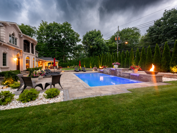 Nighttime ground-level view of the pool with fire bowls lit and landscape uplighting on surrounding plantings