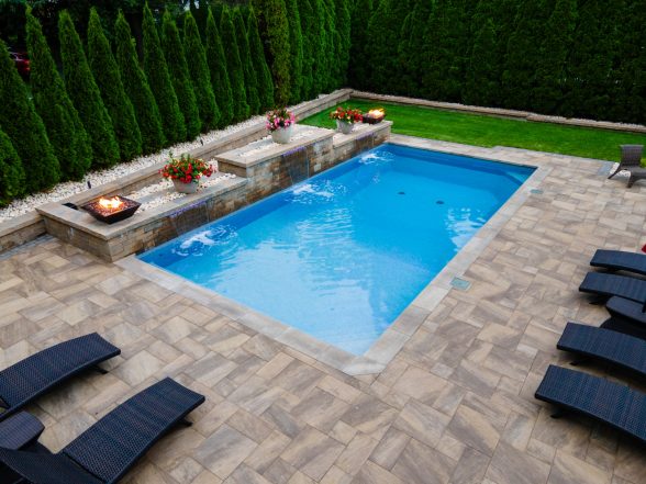 Rectangular inground pool with raised stone water feature wall, fire bowls, natural stone paver patio, and dark lounge chairs