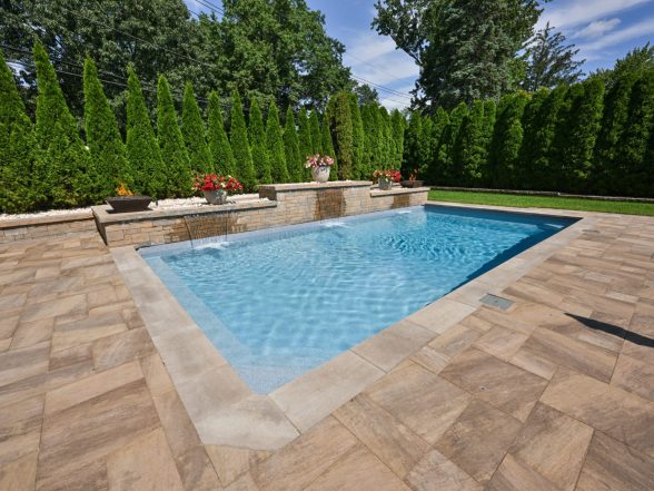 Wide ground-level view of the rectangular pool with stone paver patio, fire bowl columns, and arborvitae backdrop