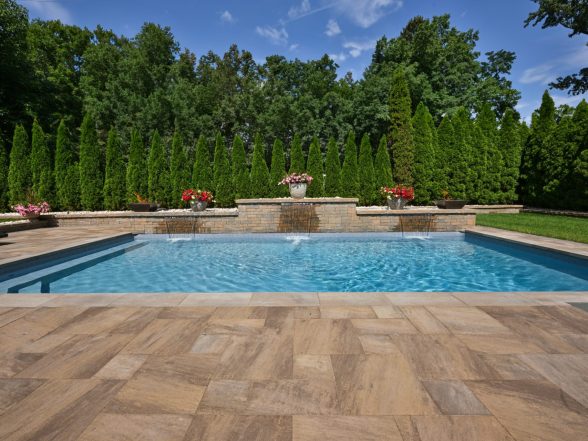 End-on view of the pool highlighting the natural stone paver pattern, raised water feature, and mature evergreen screening
