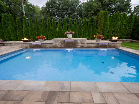 Wide view of the pool with raised sheer descent water feature, fire bowls on stone pillars, and columnar arborvitae backdrop