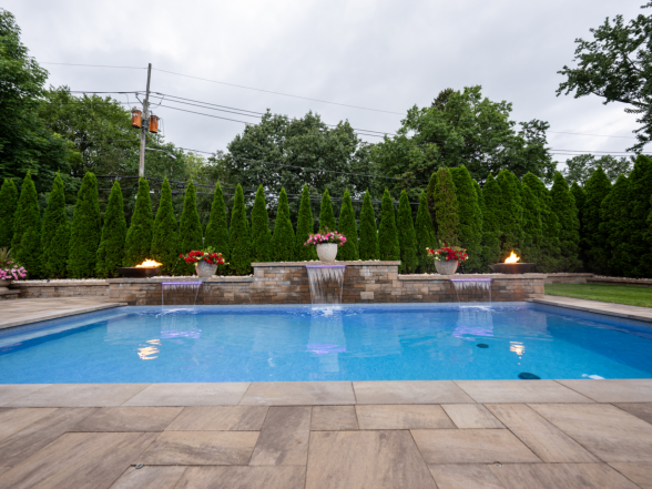 Side angle of the pool at dusk with fire bowls glowing, sheer descent waterfalls, and arborvitae screening