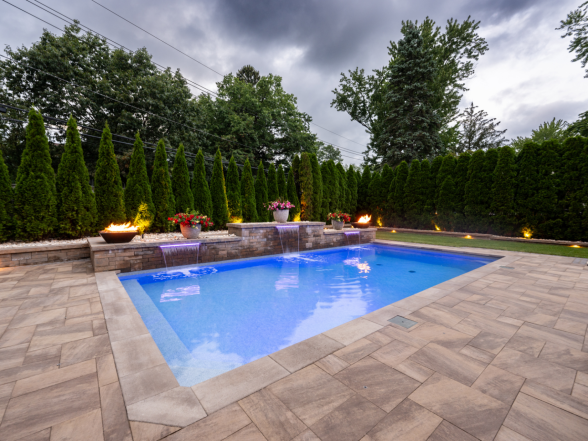Daytime view of the rectangular pool from the shallow end with natural stone paver deck and fire bowl pillars