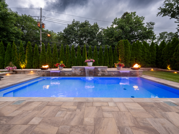 Full-length view of the pool with stone water feature wall, fire bowls, and mature landscaping under overcast skies