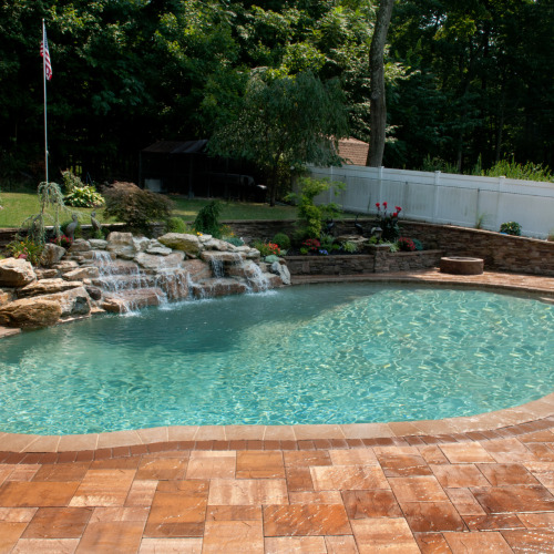 Freeform inground pool with multi-tier natural boulder waterfall, stacked stone retaining wall, brick paver patio, and colorful perennial plantings