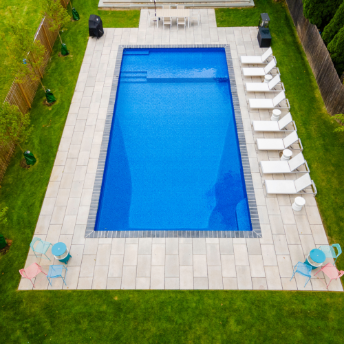 Modern rectangular swimming pool with blue water, surrounded by a spacious patio featuring white lounge chairs and colorful outdoor furniture, showcasing a completed pool project by EverClear Pools & Spas.