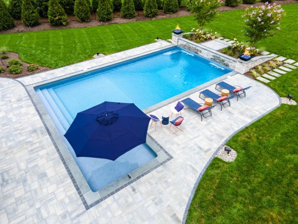 Rectangular inground pool with tanning ledge, navy patio umbrella, colorful lounge chairs, and curved paver walkway