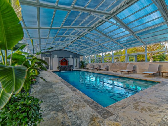 Indoor pool with clear blue water, surrounded by lounge chairs and tropical plants, featuring a modern glass roof, showcasing a completed project by EverClear Pools & Spas in Kendall Park, NJ.