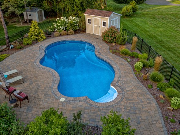Aerial view of a freeform pool with expansive paver patio, perennial landscape beds, garden shed, and aluminum pool fencing