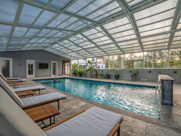 Exterior angle of the enclosed gunite pool showing the glass roof structure, travertine patio, and outdoor lounge seating