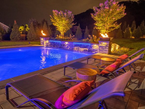 Nighttime poolside view from the patio seating area showing blue LED lighting, fire bowls, and outdoor lounge furniture