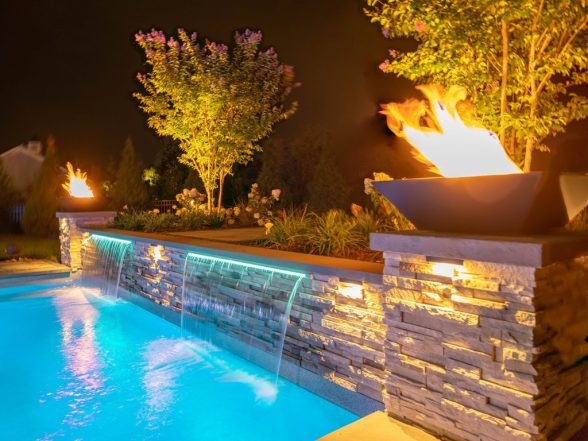 Close-up of a natural gas fire bowl on a stone pillar beside the illuminated pool at night