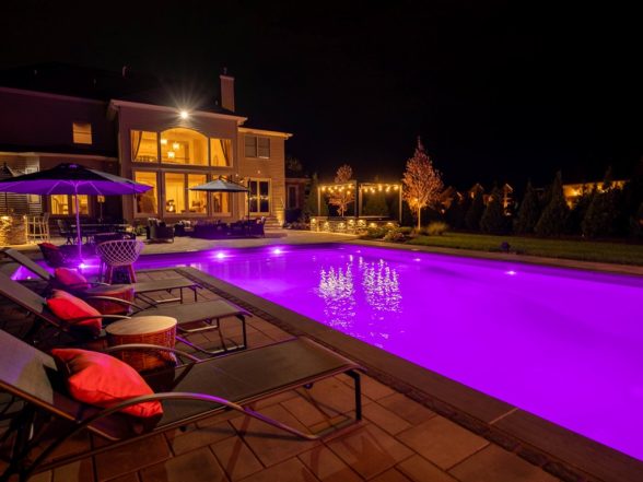 Ground-level nighttime view of the pool glowing purple with landscape uplighting on surrounding trees