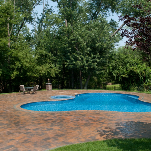 Gunite inground pool with a spa, surrounded by a brick patio and lush greenery, showcasing a completed pool project by EverClear Pools & Spas in Franklin Lakes, NJ.