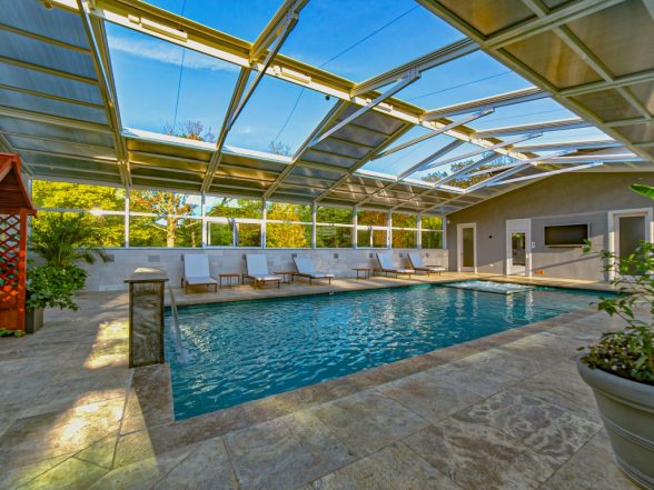 Side angle inside the pool enclosure highlighting the travertine patio, lounge seating, and glass panel roof structure