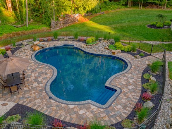 Gunite pool surrounded by a stone patio, landscaped with plants and rocks, featuring a shaded seating area, showcasing a completed project by EverClear Pools & Spas in Long Valley, NJ.