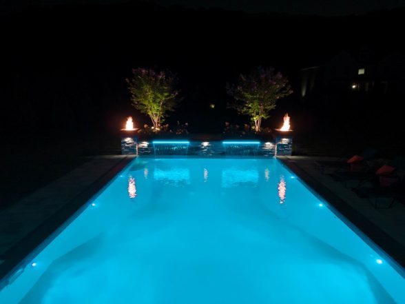 Wide-angle nighttime shot of the pool with purple LED lighting, fire bowls, and professional landscape lighting throughout the yard