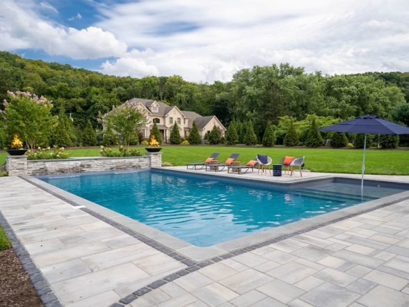 Daytime view of the rectangular inground pool with bluestone coping, Techo Bloc paver patio, and raised stone water feature pillars