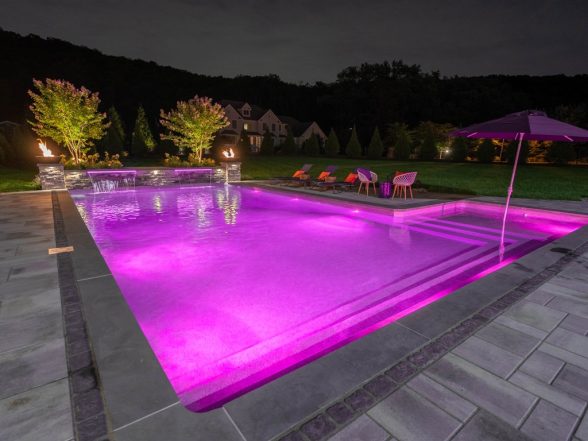 Magenta LED pool lighting at night with raised stone columns, sheer descent waterfalls, and patio umbrella