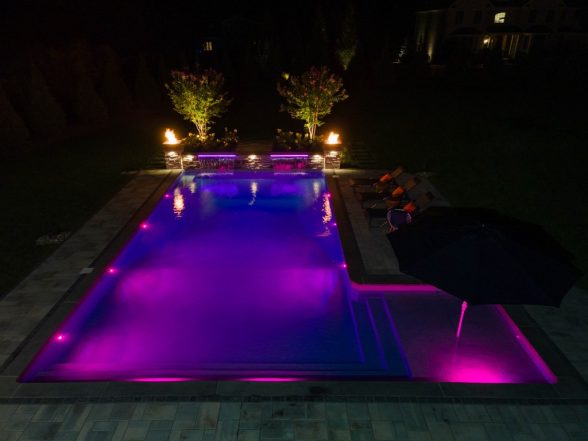 Nighttime aerial of the pool illuminated with purple LED lighting, showing raised stone water feature pillars and fire bowls