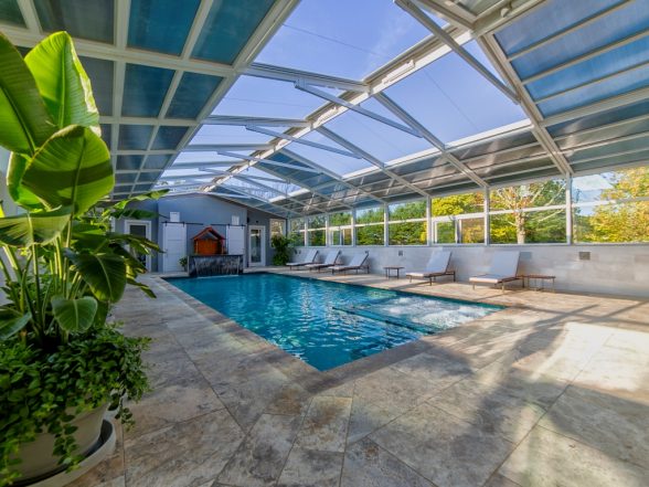 End-on perspective of the enclosed pool with glass roof panels, natural stone decking, and tropical landscaping