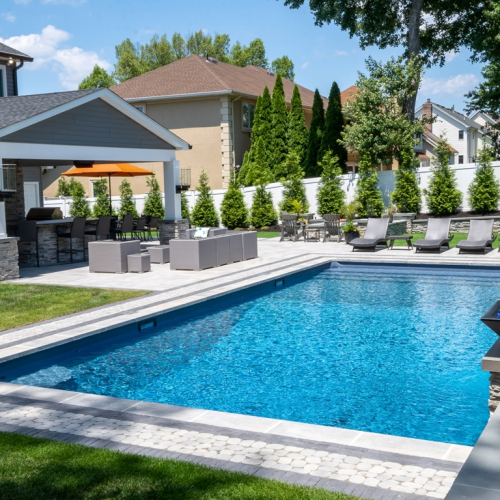 Modern backyard swimming pool with lounge chairs, fire feature, and covered patio, showcasing a completed project by EverClear Pools & Spas in Wayne, NJ.