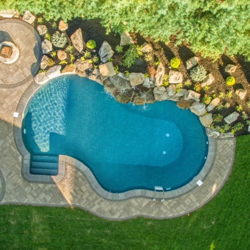 Aerial view of a beautifully designed vinyl pool surrounded by landscaped stones, greenery, and a paved patio with lounge chairs, showcasing a completed pool project by EverClear Pools & Spas in Fairfield, NJ.