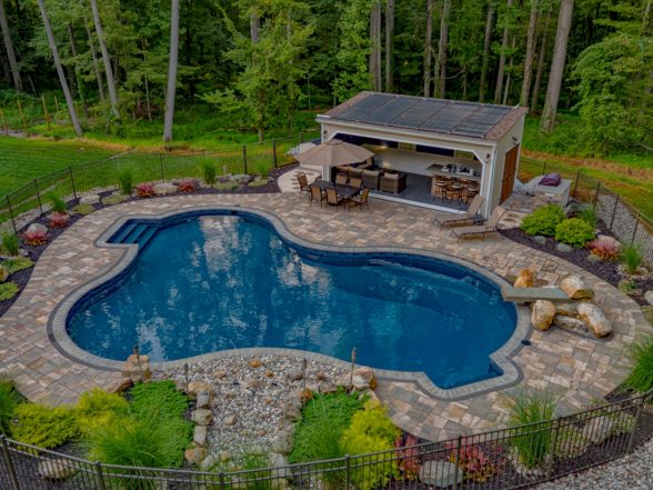 Aerial view of a freeform inground pool with paver patio, outdoor kitchen pavilion with bar seating, boulder retaining wall landscaping, and aluminum pool fence