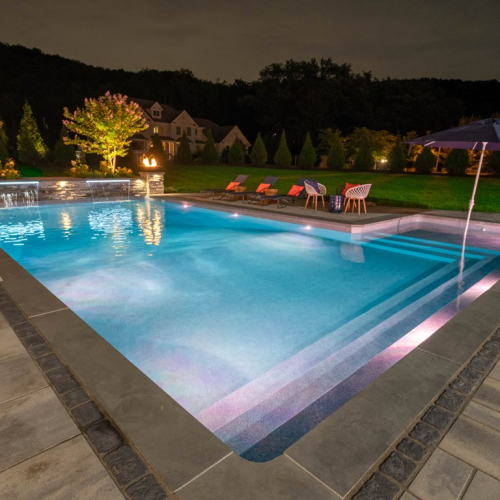 Illuminated in evening light, a modern rectangular pool with cascading water features, surrounded by lush greenery and stylish outdoor seating, showcasing EverClear Pools & Spas elegant pool design and maintenance in Denville, NJ.